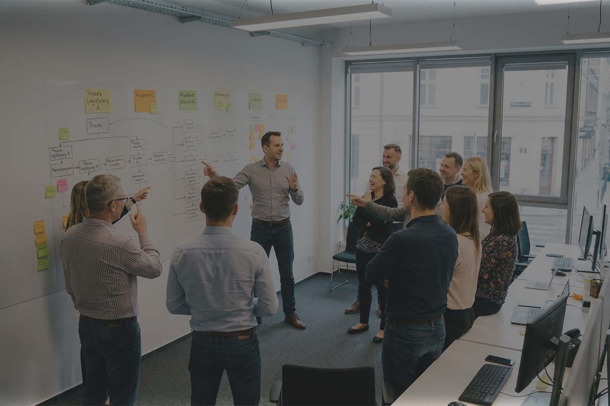 Szeroki kadr nowoczesnego biura: zespół pracowników przy tablicy z mapą procesów i napisem Wyzwania, trener facylituje dyskusję.
