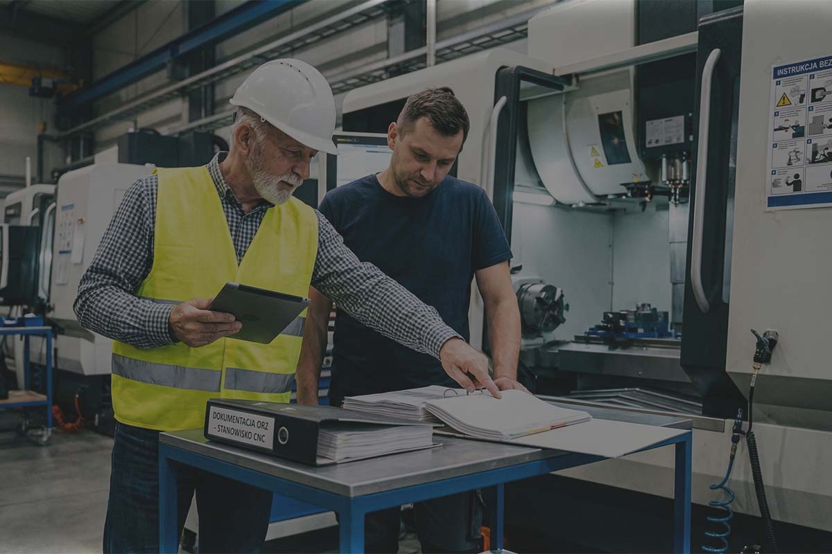 Audytor na hali omawia ocenę ryzyka przy segregatorze z napisem "DOKUMENTACJA ORZ - STANOWISKO CNC". W tle wiszą instrukcje bezpieczeństwa.
